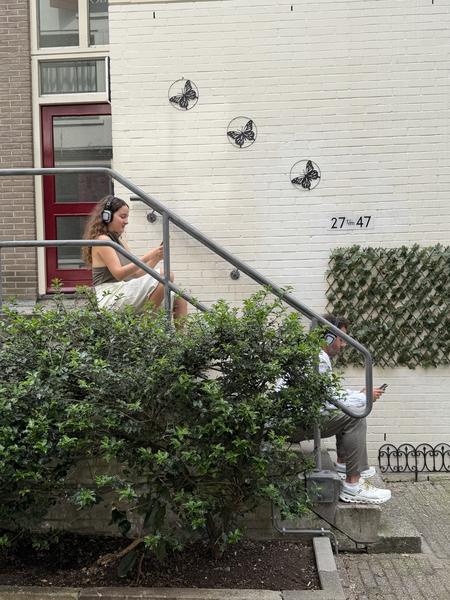 Two individuals are seen sitting on concrete steps, engrossed in their smartphones and wearing headphones, in Brandewijnsteeg, Nieuwmarktbuurt, Amsterdam